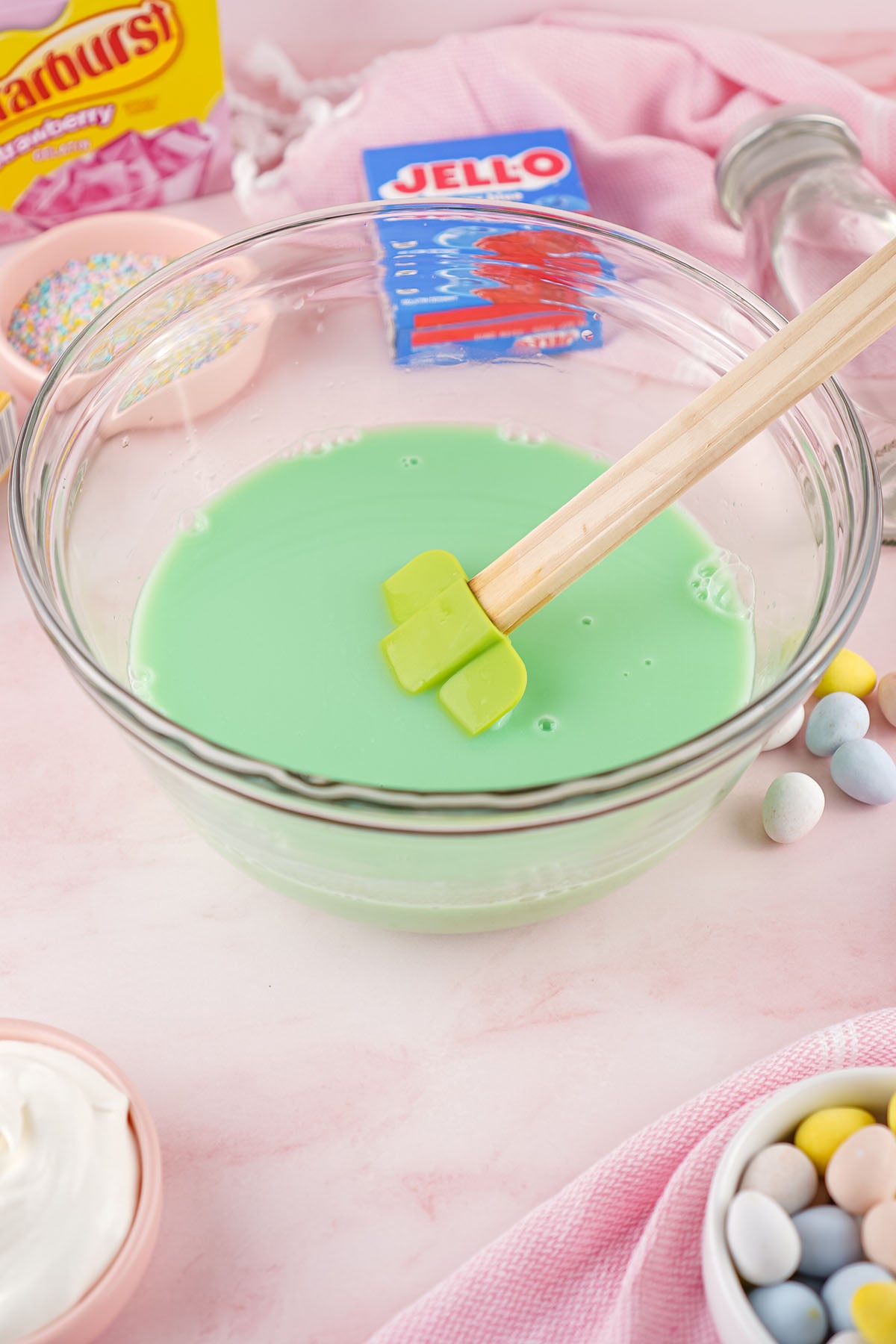 A glass mixing bowl containing creamy lime-green Jello being stirred with a green spatula, surrounded by Easter candy and boxes of gelatin.