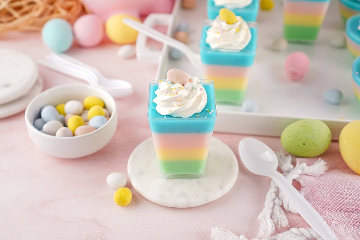a layered pastel jello cup on a small marble coaster, surrounded by white spoons, a bowl of candy eggs, and more dessert cups in the background.
