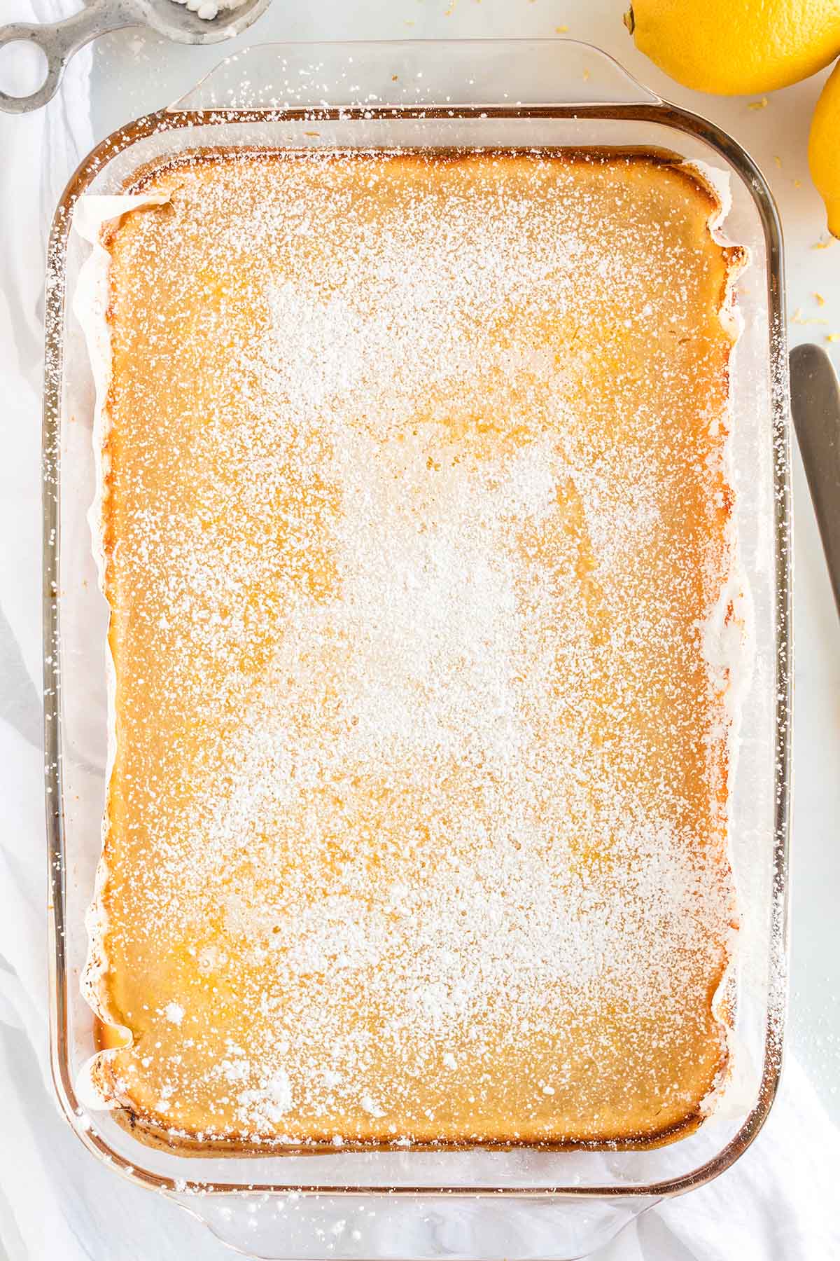 Freshly baked lemon bars in a glass pan, topped with a generous dusting of powdered sugar.