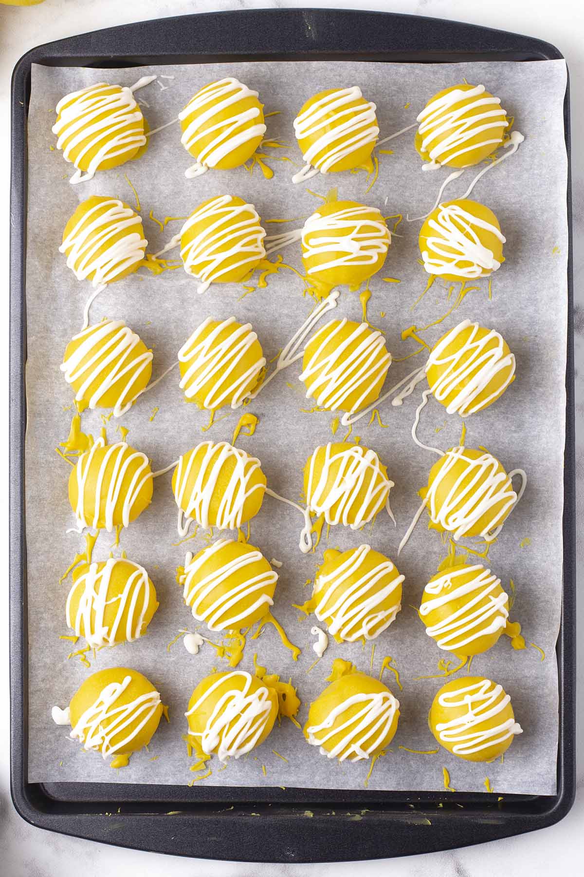 Lemon cheesecake bites coated in yellow candy and drizzled with white chocolate on a parchment-lined baking sheet.