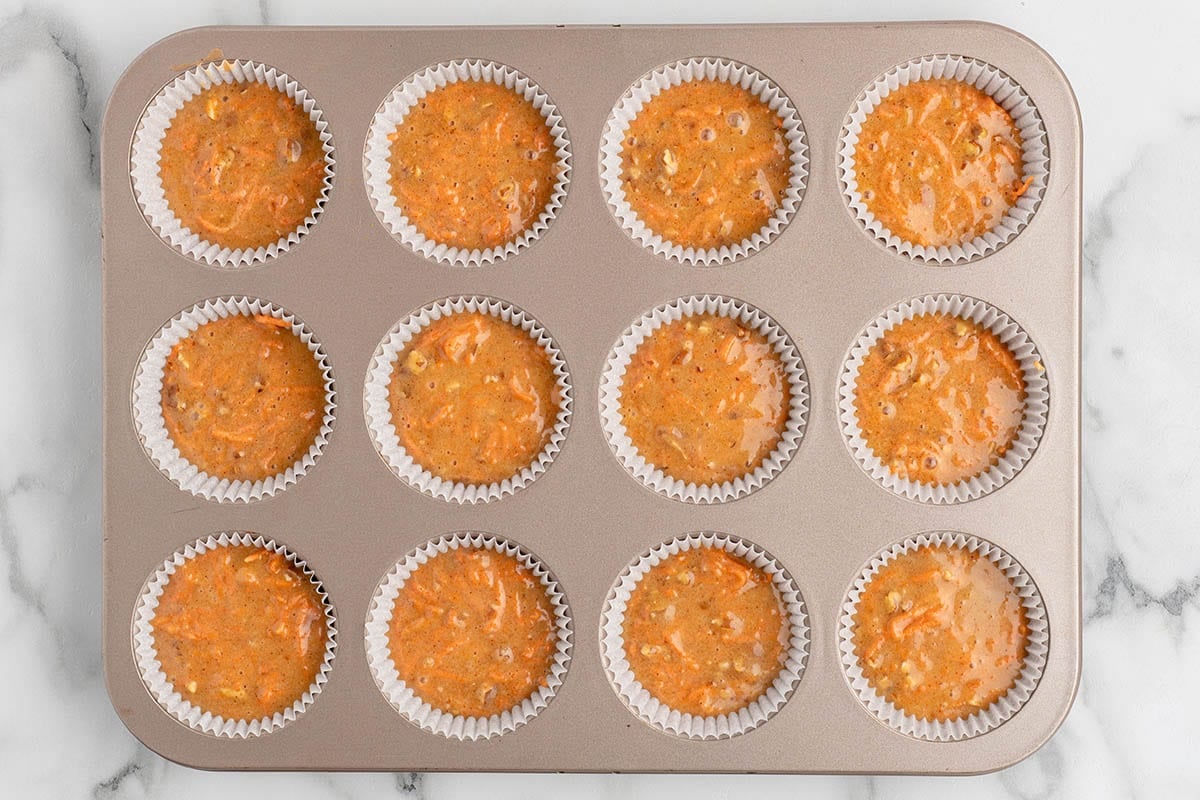 a 12-count muffin tin with white liners filled about three-quarters full with spiced carrot cake batter, showing visible shreds of carrot and chopped pecans.