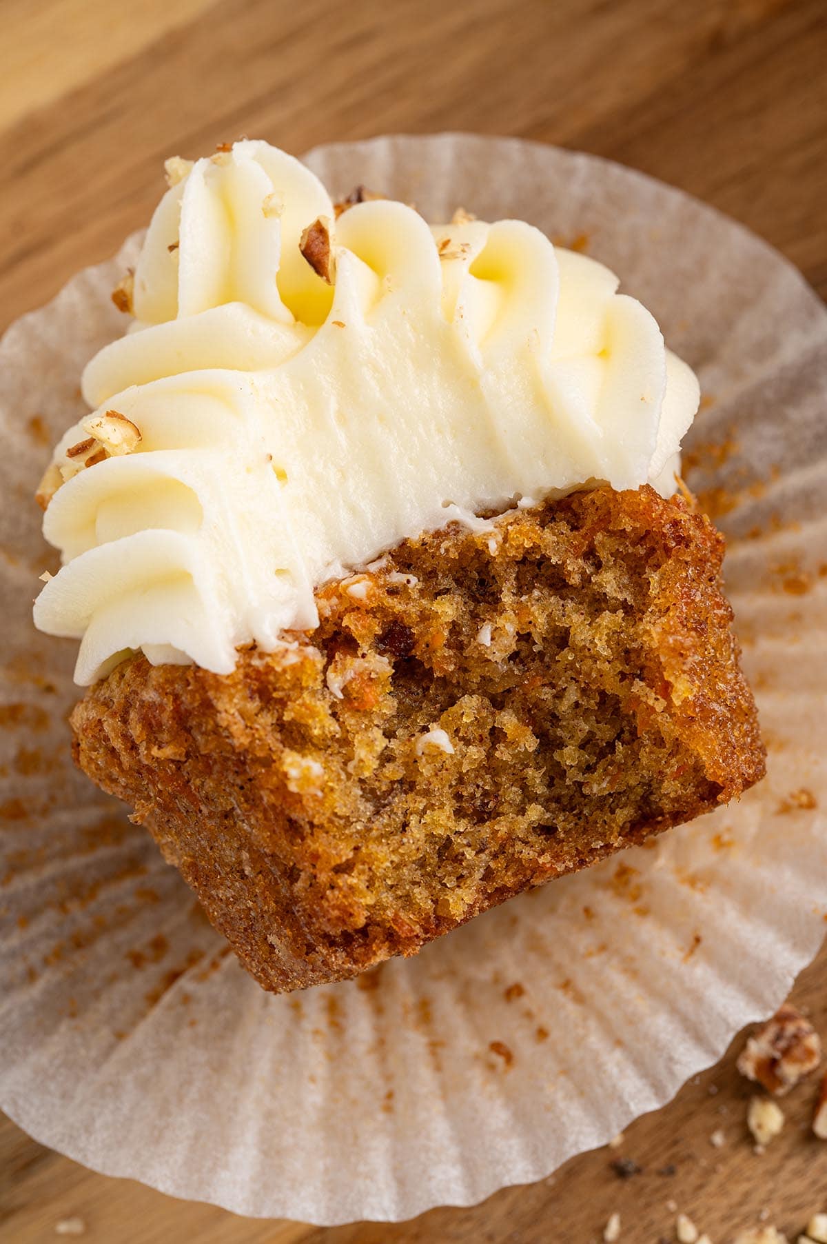 a carrot cake cupcake with the wrapper pulled down and a bite removed, showcasing the moist spiced crumb and a thick swirl of cream cheese frosting.