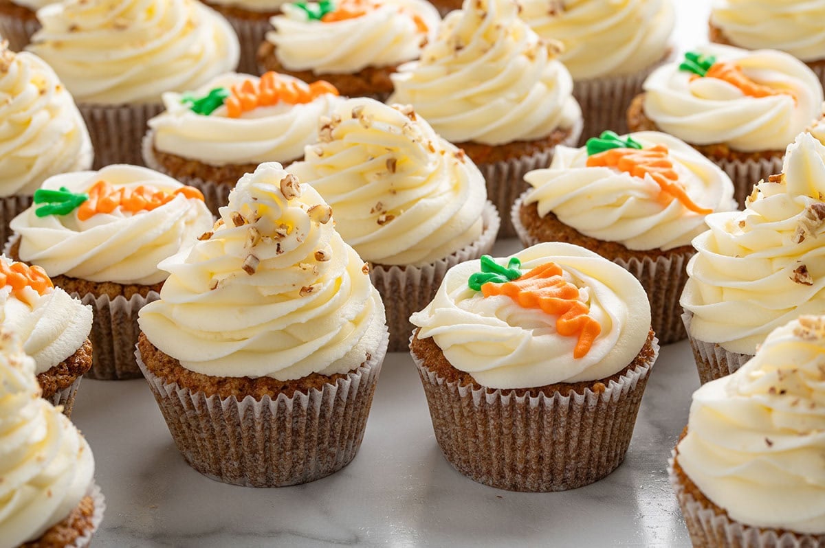 carrot cake cupcakes on a marble surface, featuring swirls of cream cheese frosting decorated with either chopped pecans or orange and green piped frosting carrots.