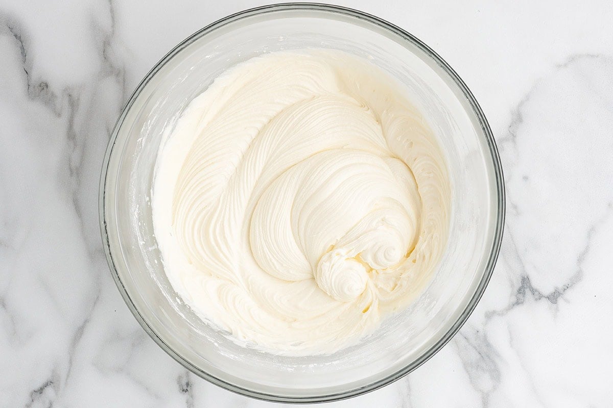 a glass bowl filled with thick, creamy white cream cheese frosting, showing smooth swirls from being whipped.