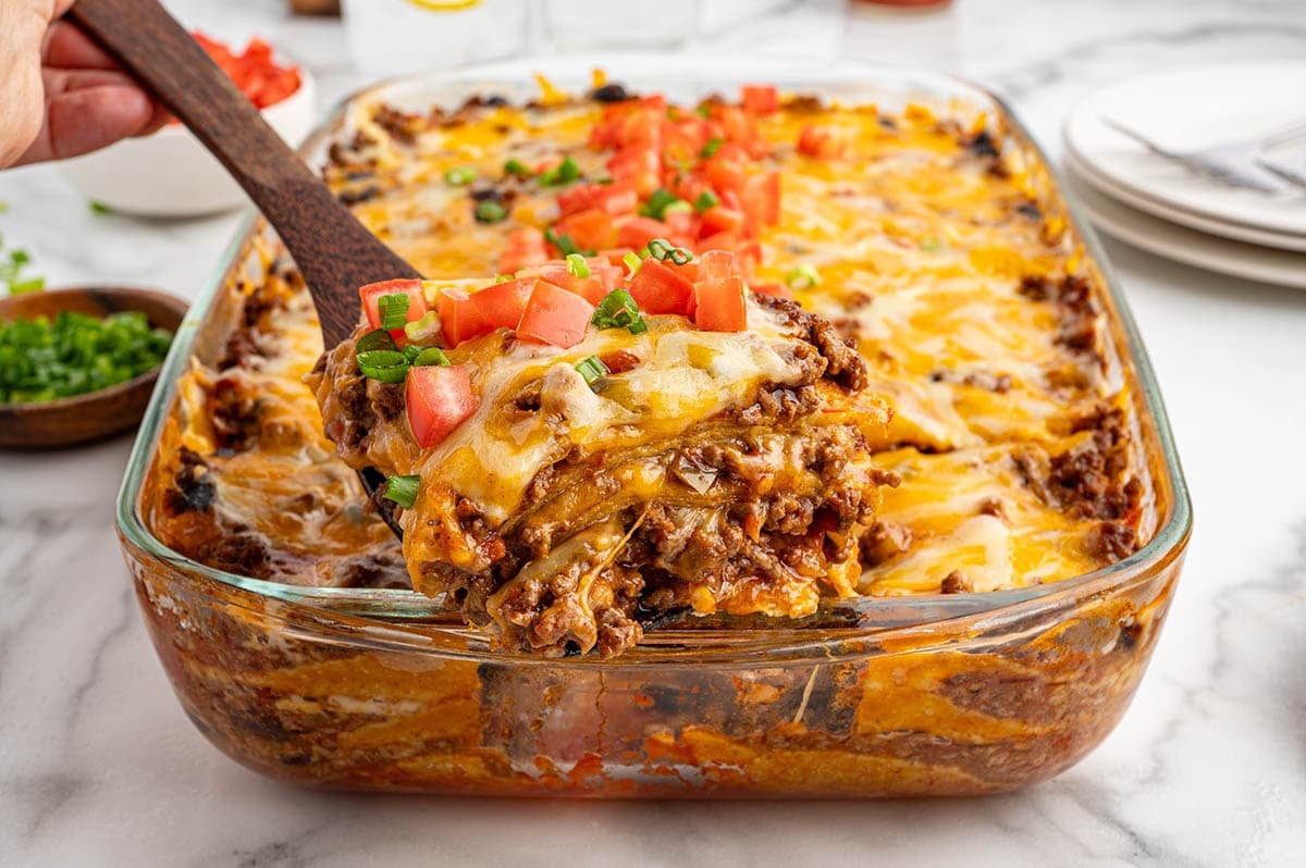 A wooden spatula lifting a cheesy serving of ground beef Mexican casserole from a glass baking dish.