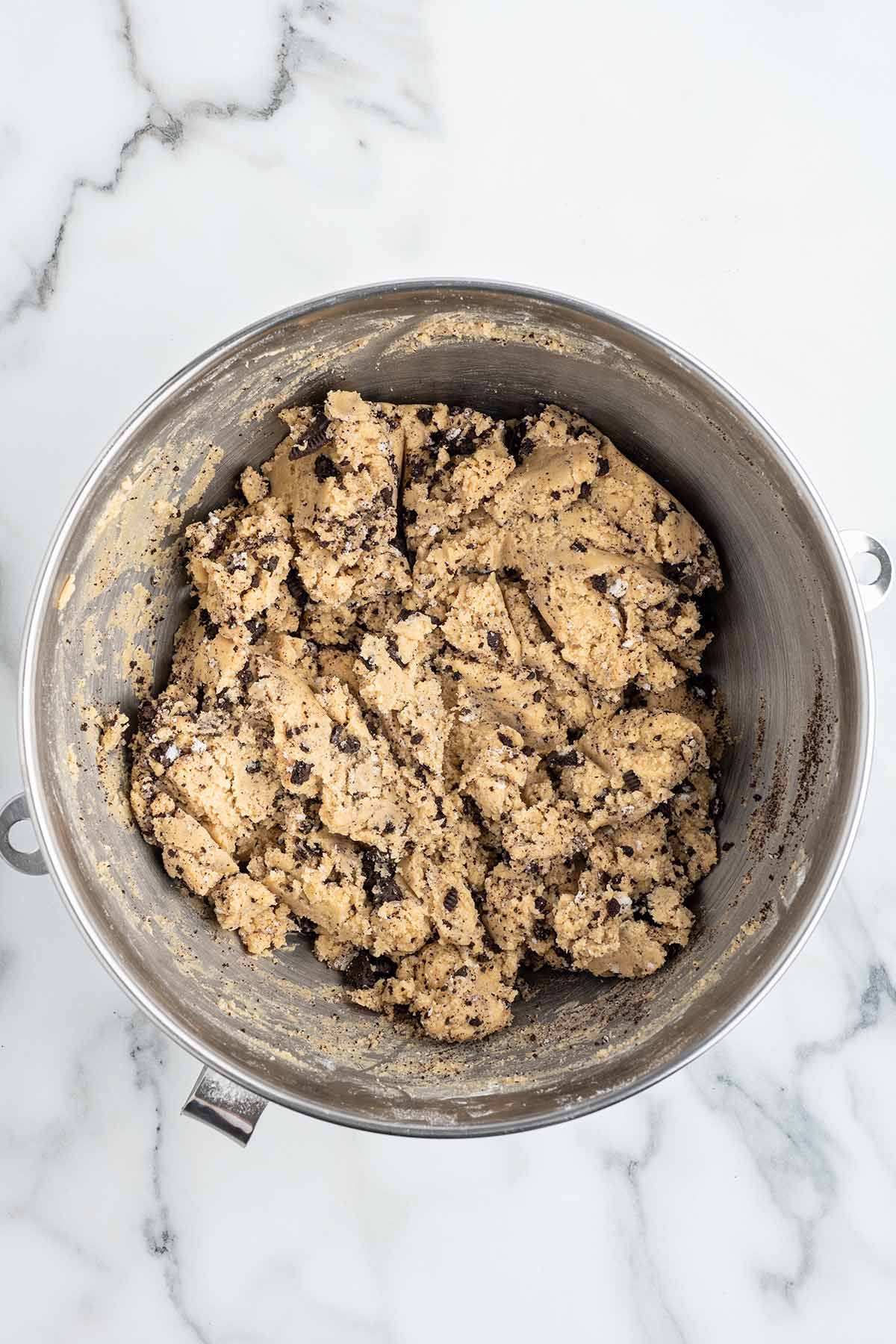 A large metal mixing bowl containing thick cookie dough mixed with dark chocolate cookie chunks.