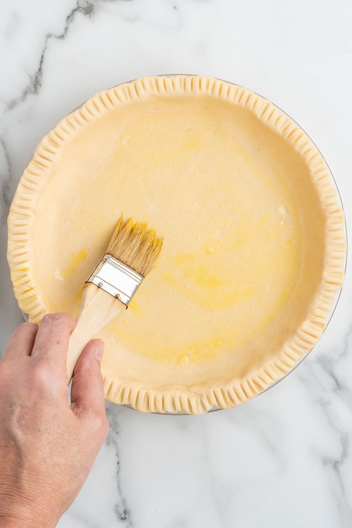 A hand using a pastry brush to apply a golden egg wash to the bottom of a raw pie crust in a glass dish.