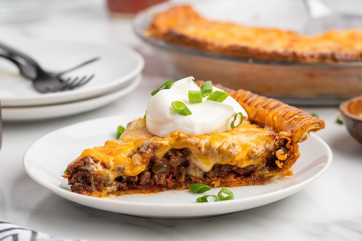 A side view slice of taco pie with a golden pastry crust, savory meat filling, and melted cheese, topped with sour cream and green onions.