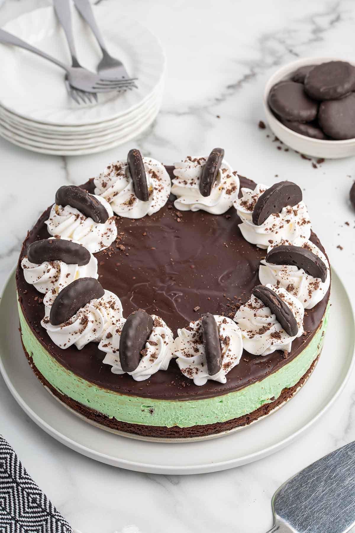A whole mint green cheesecake on a white plate, topped with chocolate ganache and a ring of whipped cream swirls garnished with Thin Mint cookies.