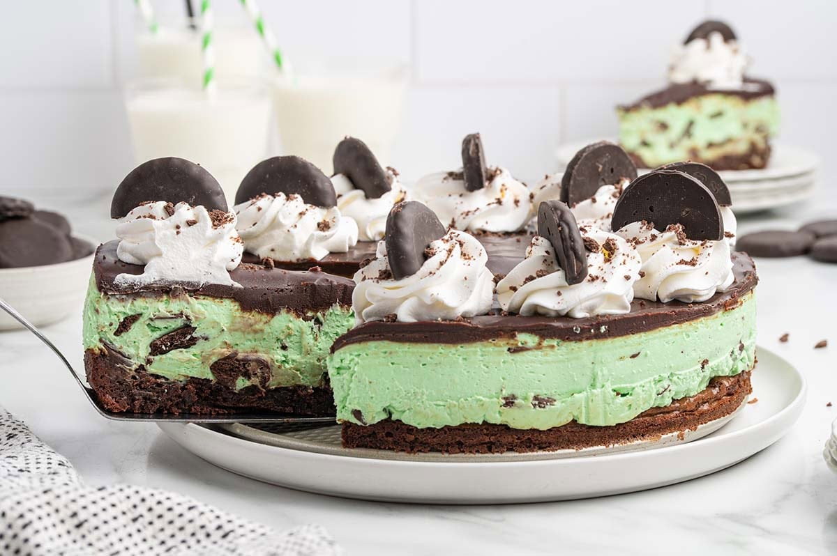 A whole mint green cheesecake topped with chocolate ganache, whipped cream swirls, and Thin Mint cookies, with one slice being lifted away.