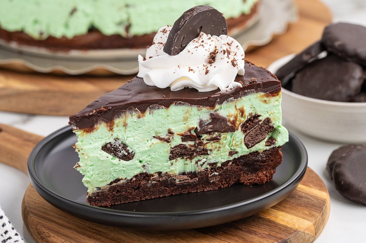 A side view slice of mint green cheesecake with cookie chunks, topped with dark chocolate and whipped cream.