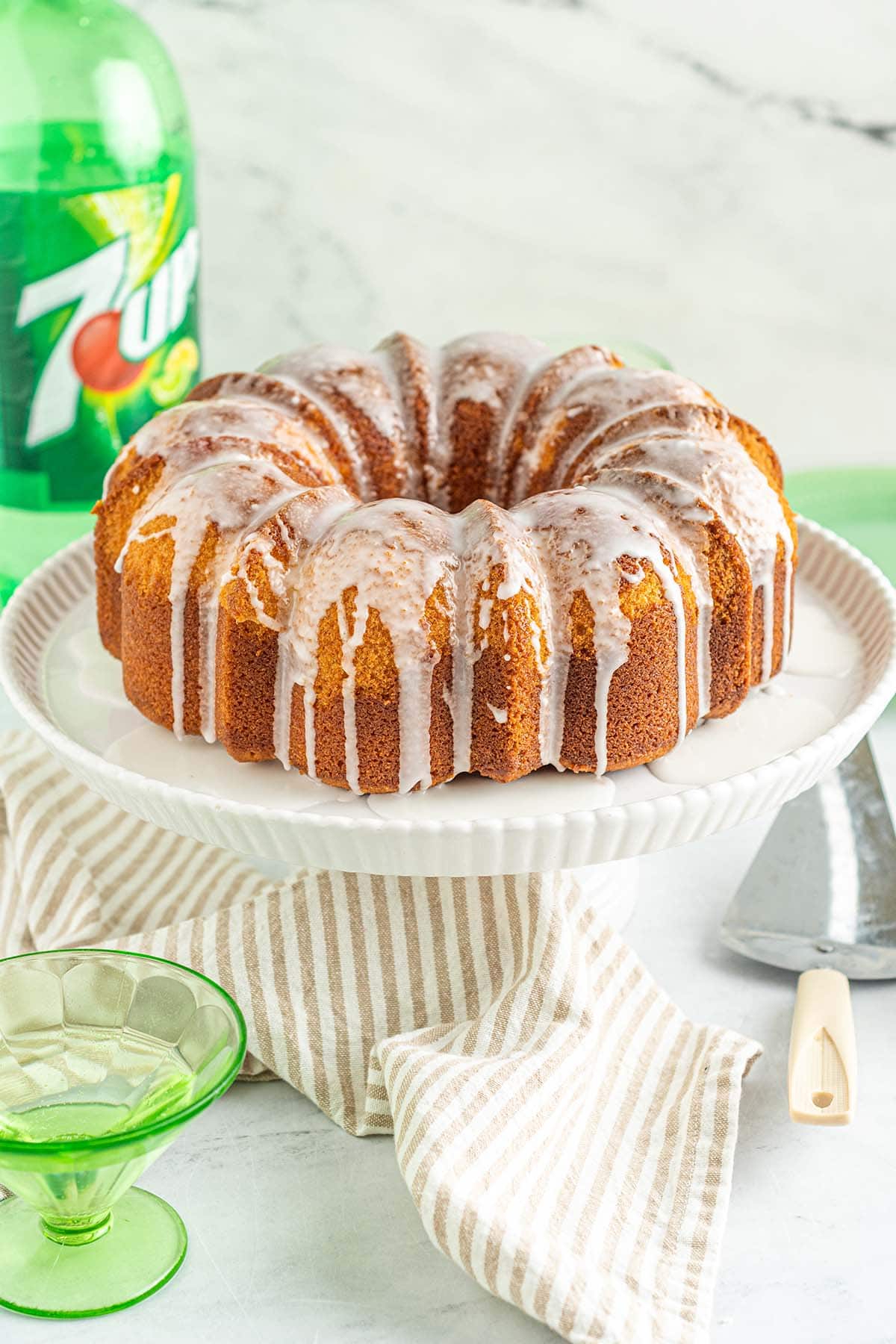 7Up pound cake with white glaze drizzled over the top on a white cake stand, with a bottle of 7Up in the background.