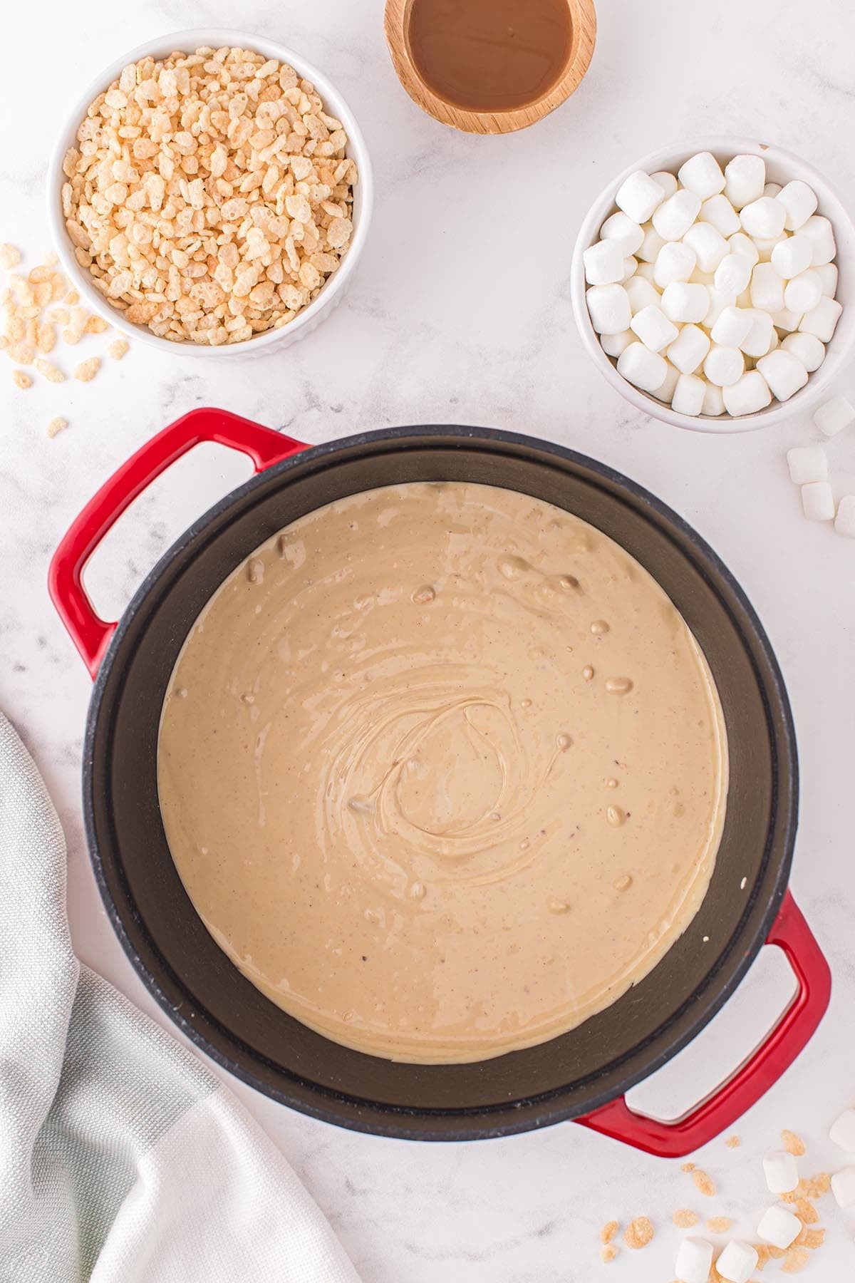 Melted white chocolate and peanut butter mixture in a red dutch oven with bowls of Rice Krispies and mini marshmallows nearby.