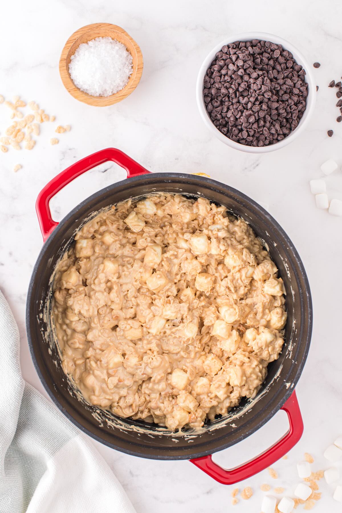 Rice Krispies and marshmallows folded into peanut butter mixture in a red dutch oven with chocolate chips and sea salt in the background.