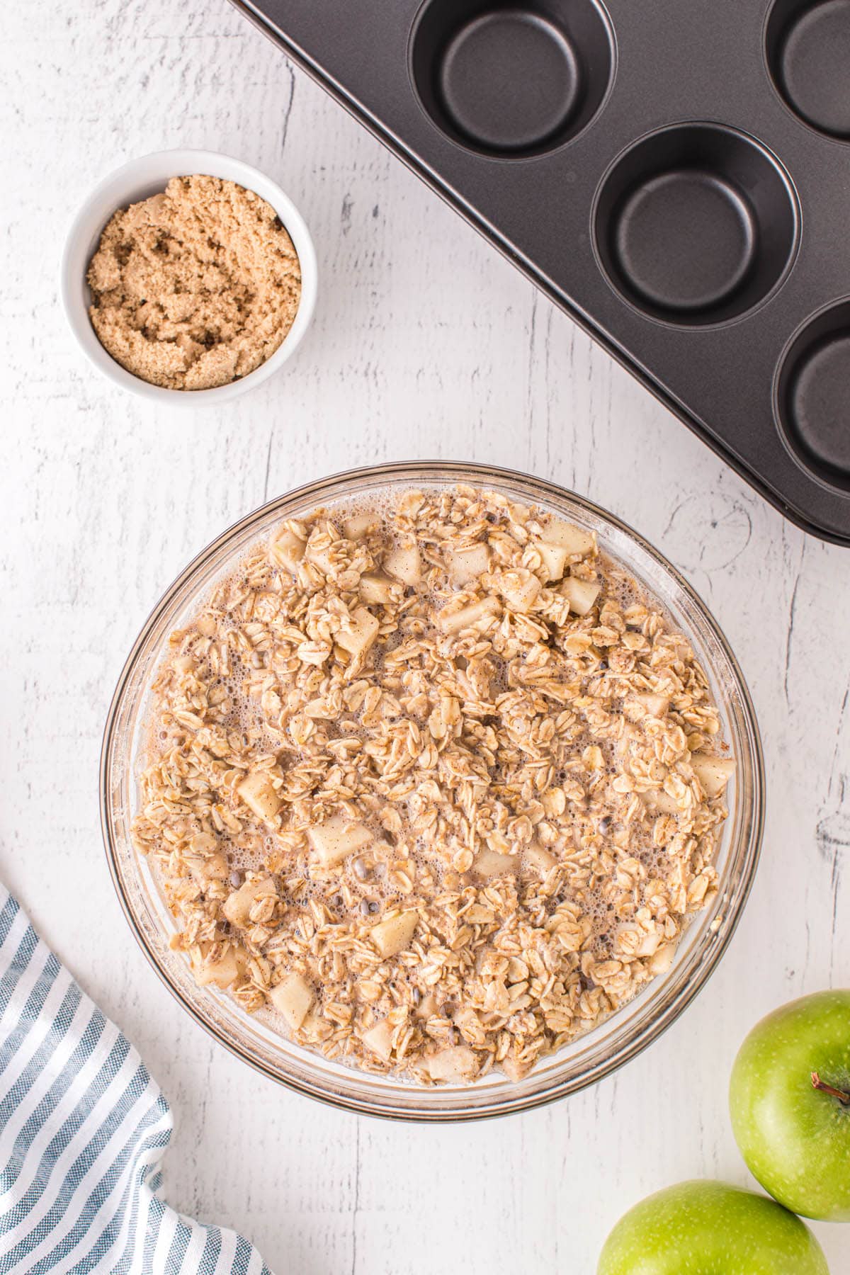 A top-down view of a bowl filled with a mixture of oats, milk, and diced apples, likely for baked oatmeal cups. Nearby are a muffin tin, a small bowl of brown sugar, and green apples on a light surface.