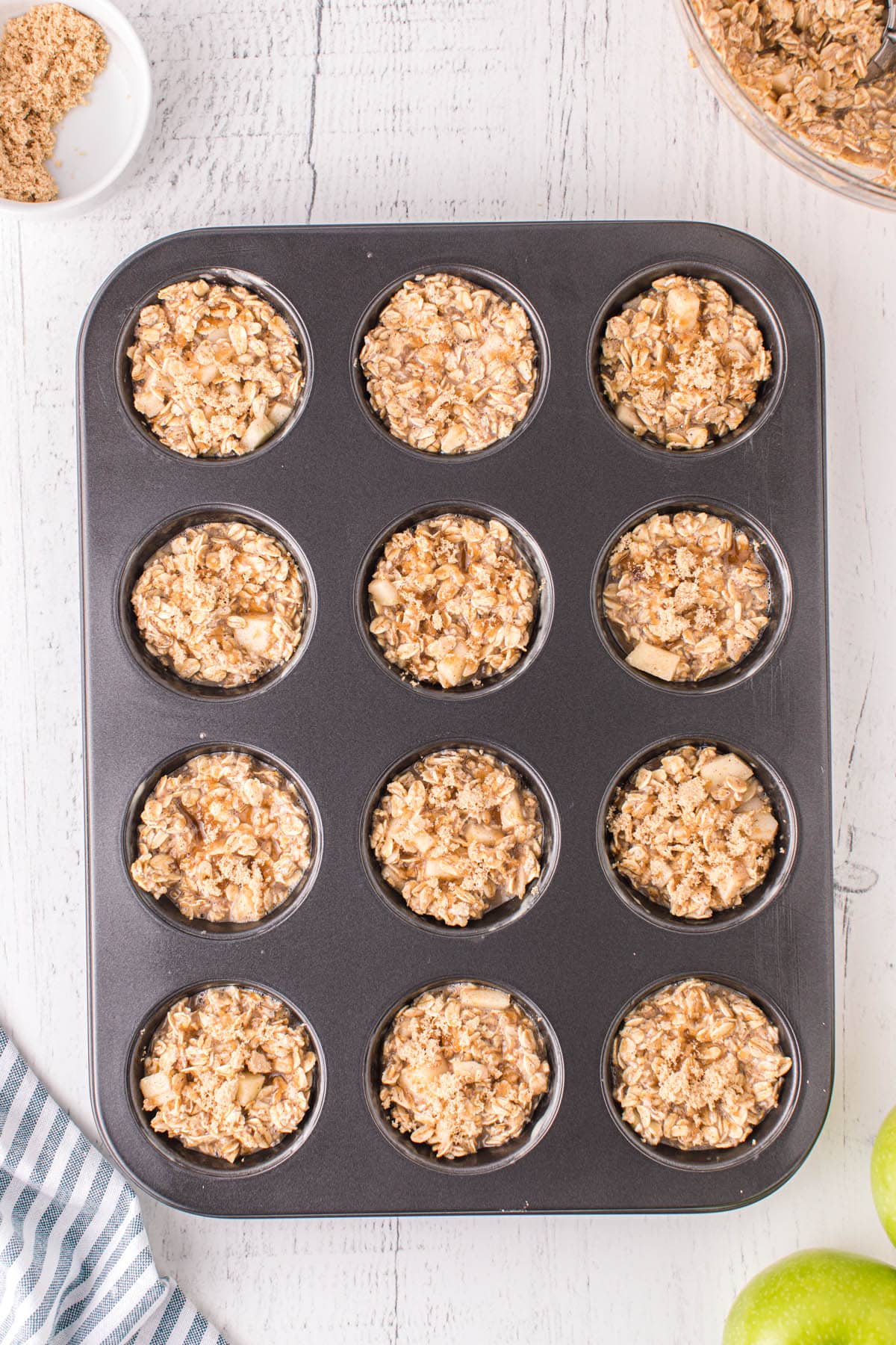 A top-down view of a muffin tin filled with portions of oat and apple mixture, ready to be baked into oatmeal cups. Brown sugar is sprinkled on top, with extra ingredients and apples visible around the tray.