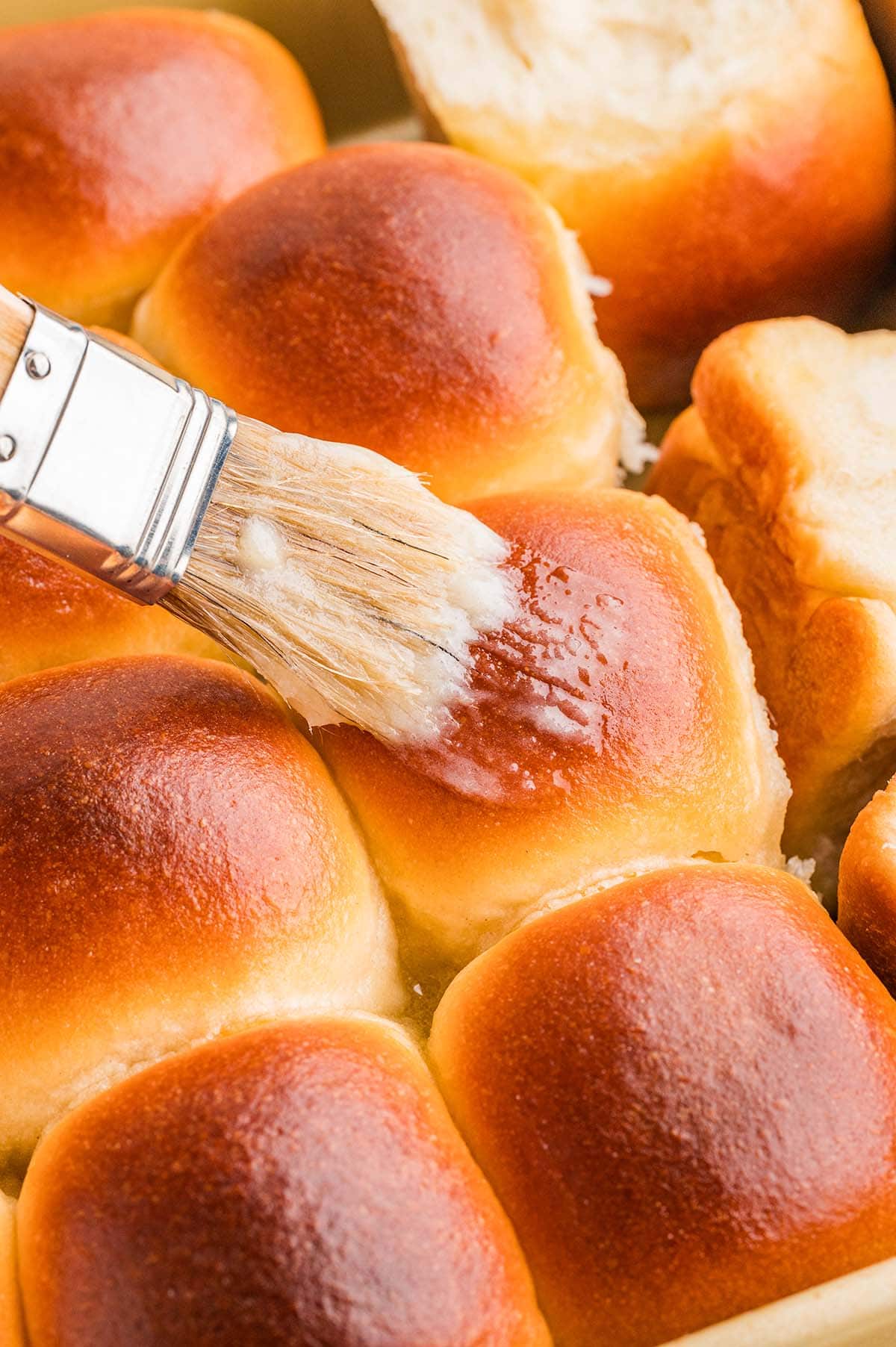 Pastry brush spreading melted butter over hot baked cafeteria rolls in a pan.