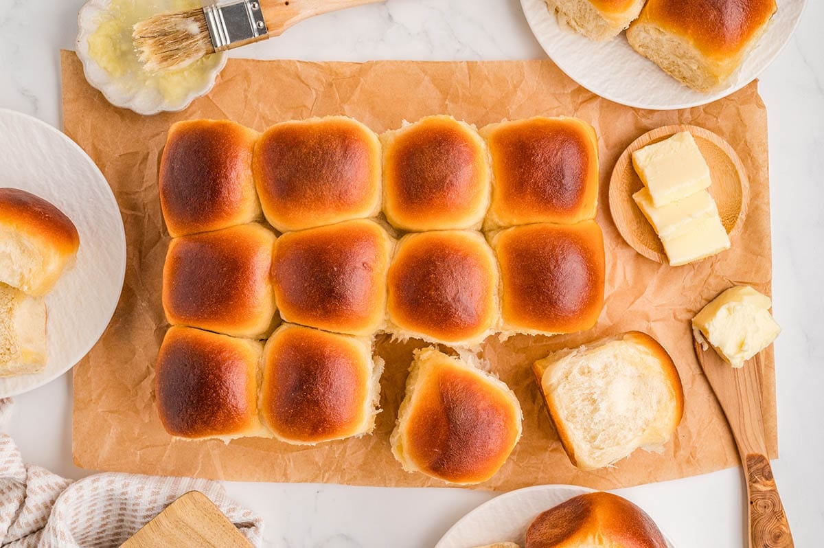 Golden brown cafeteria rolls on parchment paper with one roll pulled apart, surrounded by pats of butter and a pastry brush.