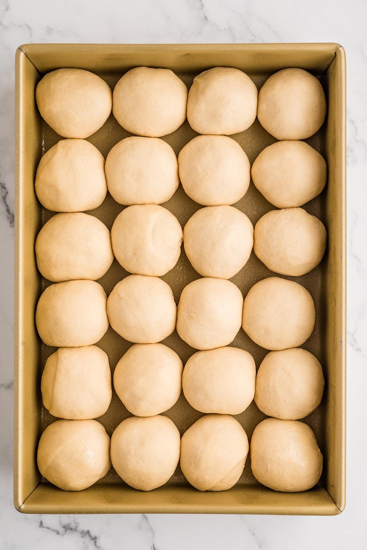 Twenty unbaked roll dough balls arranged in a gold baking pan before proofing.
