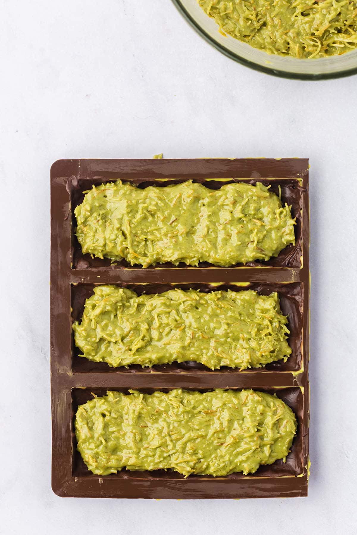  Chocolate bar mold filled with pistachio and kataifi filling, with a bowl of the mixture in the background.