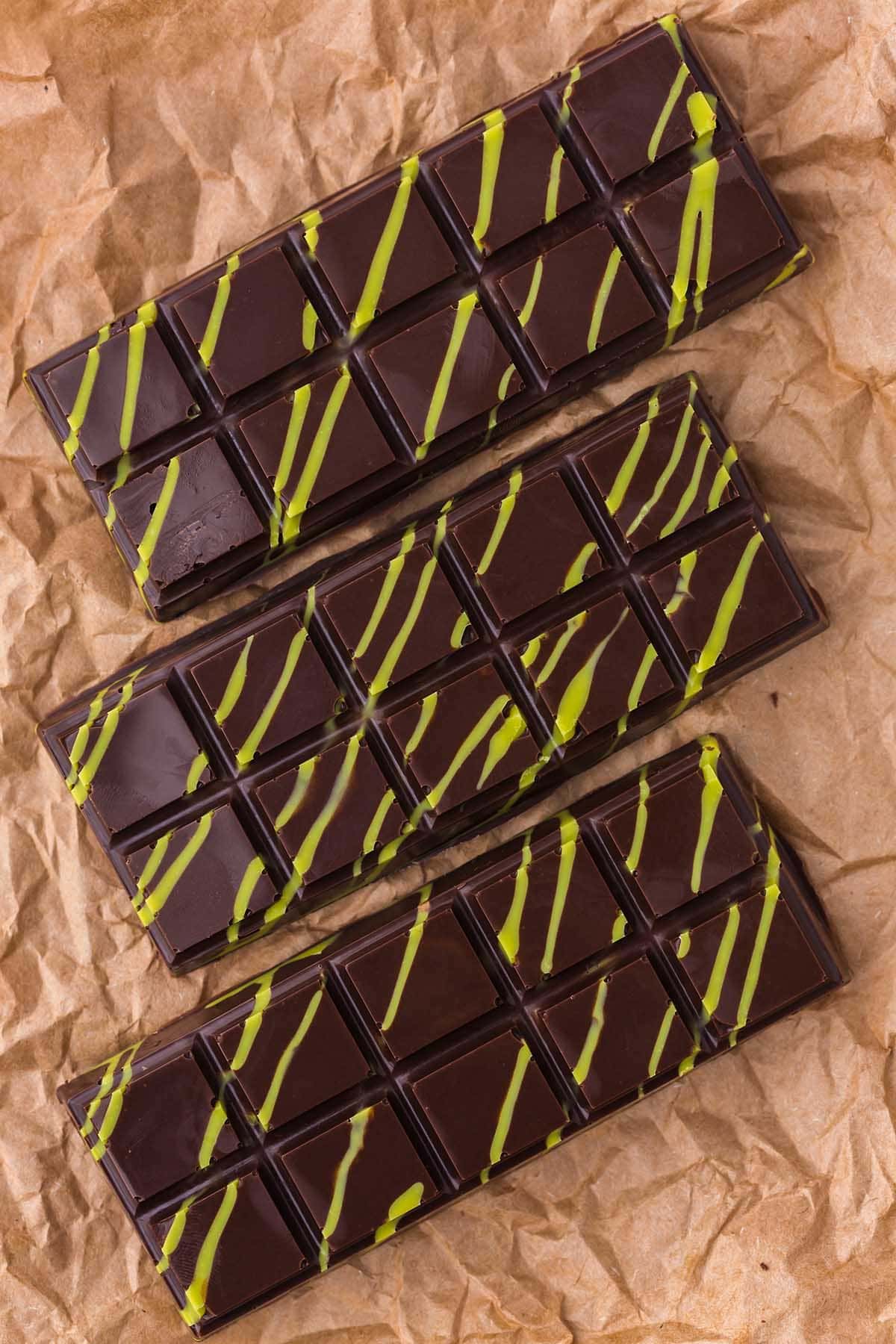 Three dark chocolate Dubai bars with green pistachio drizzle laid on crinkled brown parchment paper.