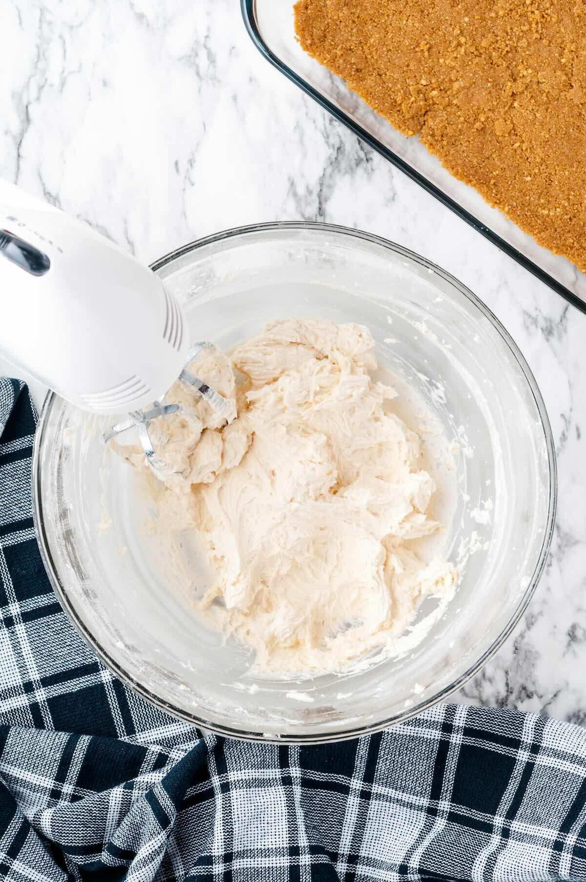 Hand mixer beating cream cheese filling in a glass bowl, with the graham cracker crust visible in the background.