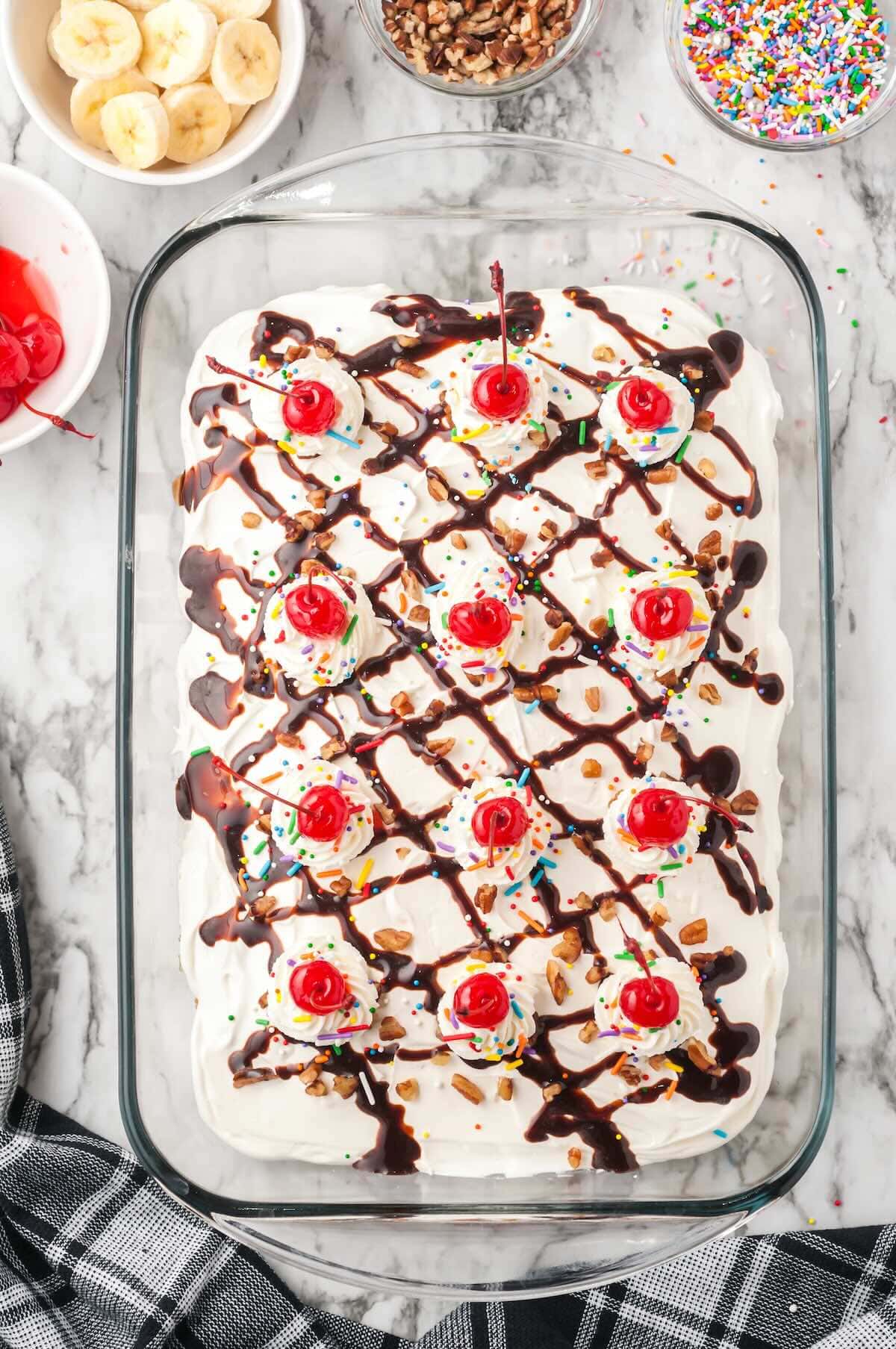 Fully assembled banana split cake topped with whipped cream swirls, maraschino cherries, chocolate sauce drizzle, sprinkles, and pecans.