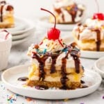 A slice of banana split cake on a white plate, topped with whipped cream, chocolate sauce, rainbow sprinkles, and a maraschino cherry.
