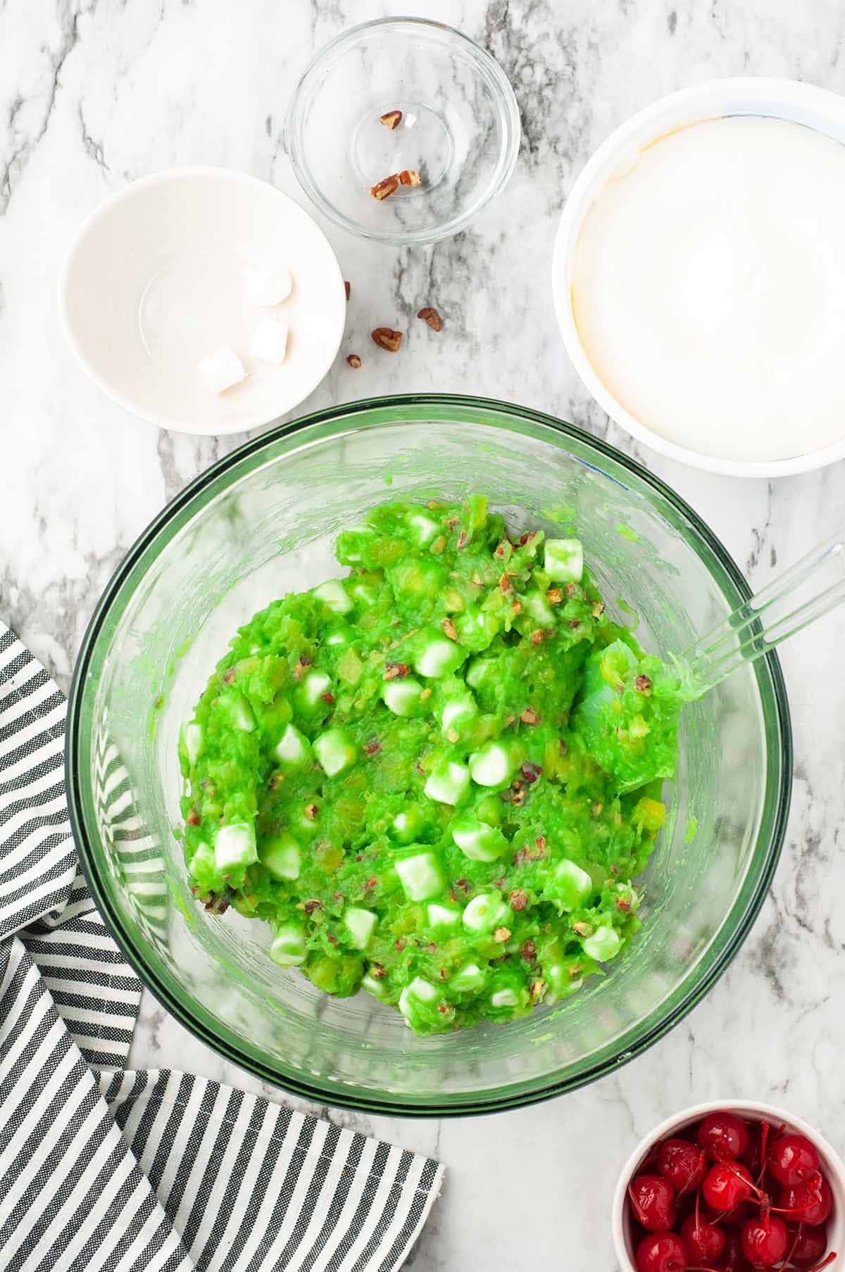 Watergate salad mixture with marshmallows and pecans folded in a glass bowl with remaining ingredients nearby.