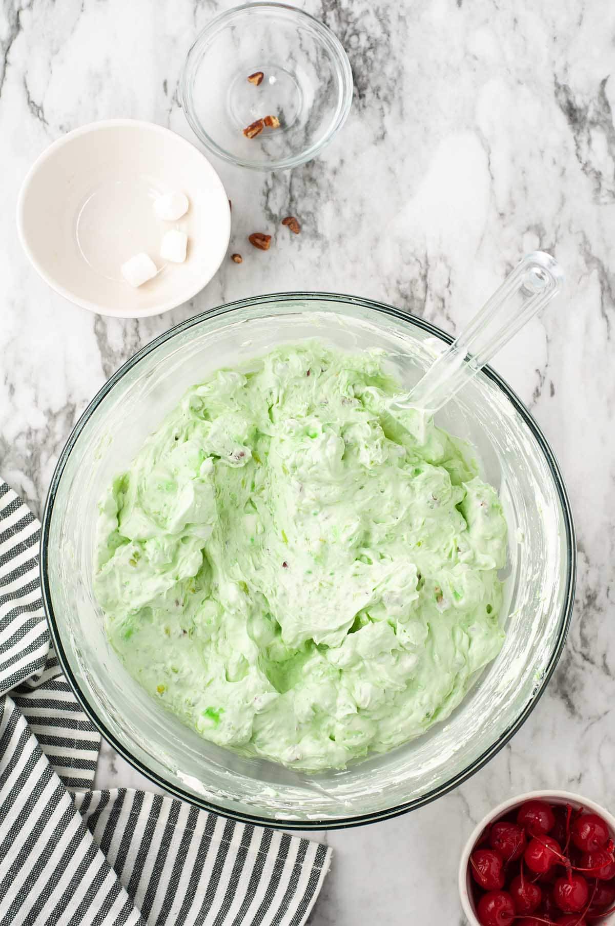 Fully mixed watergate salad in a glass bowl with whipped cream folded in next to maraschino cherries.