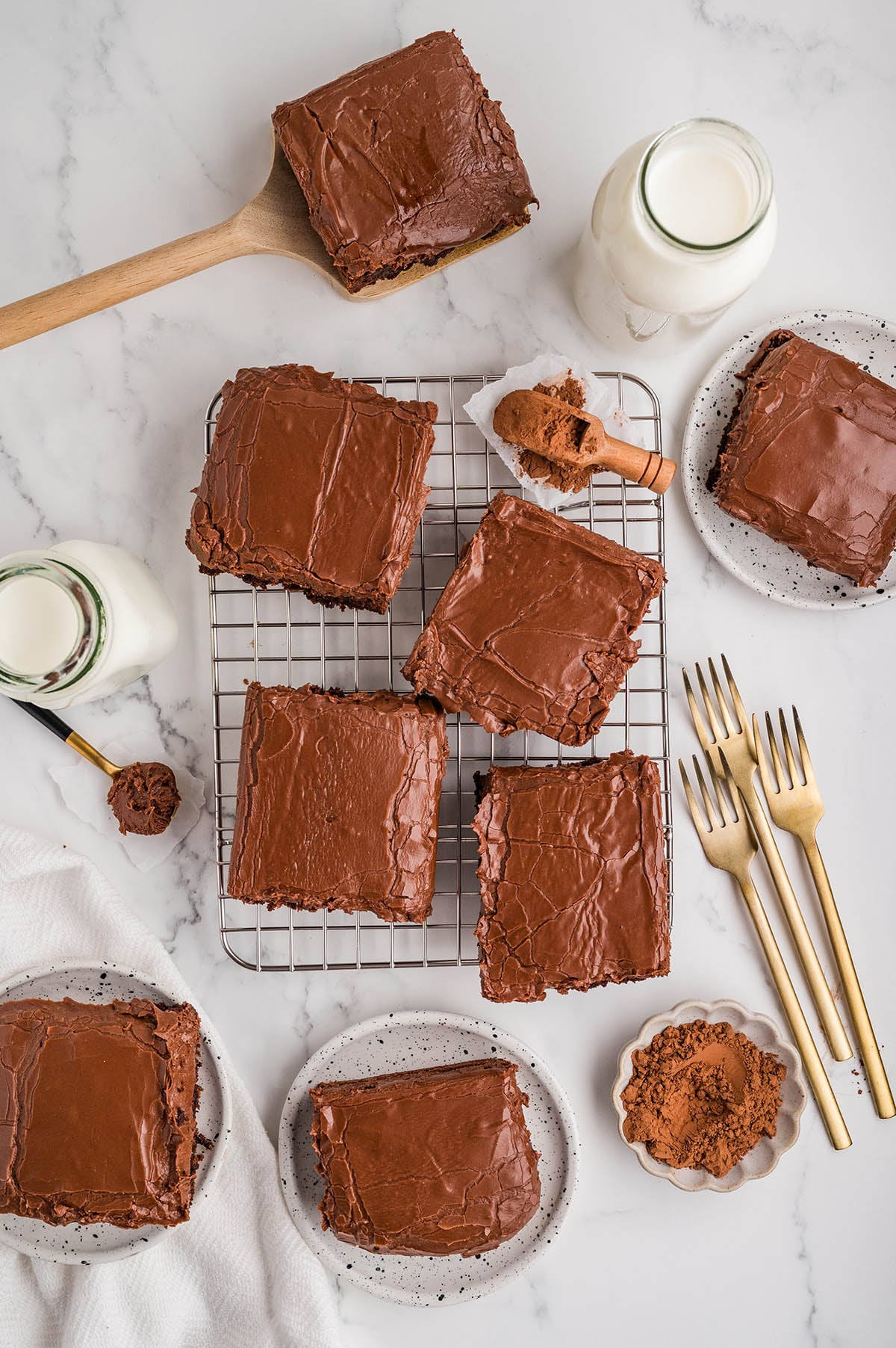 Frosted brownies arranged on a cooling rack and plates with milk and cocoa nearby.