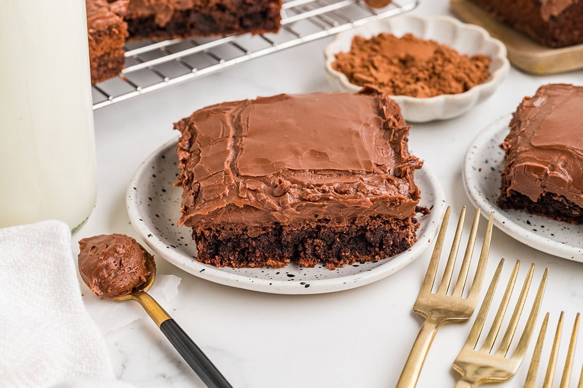 Chocolate brownie with thick frosting on a plate with milk and forks nearby.