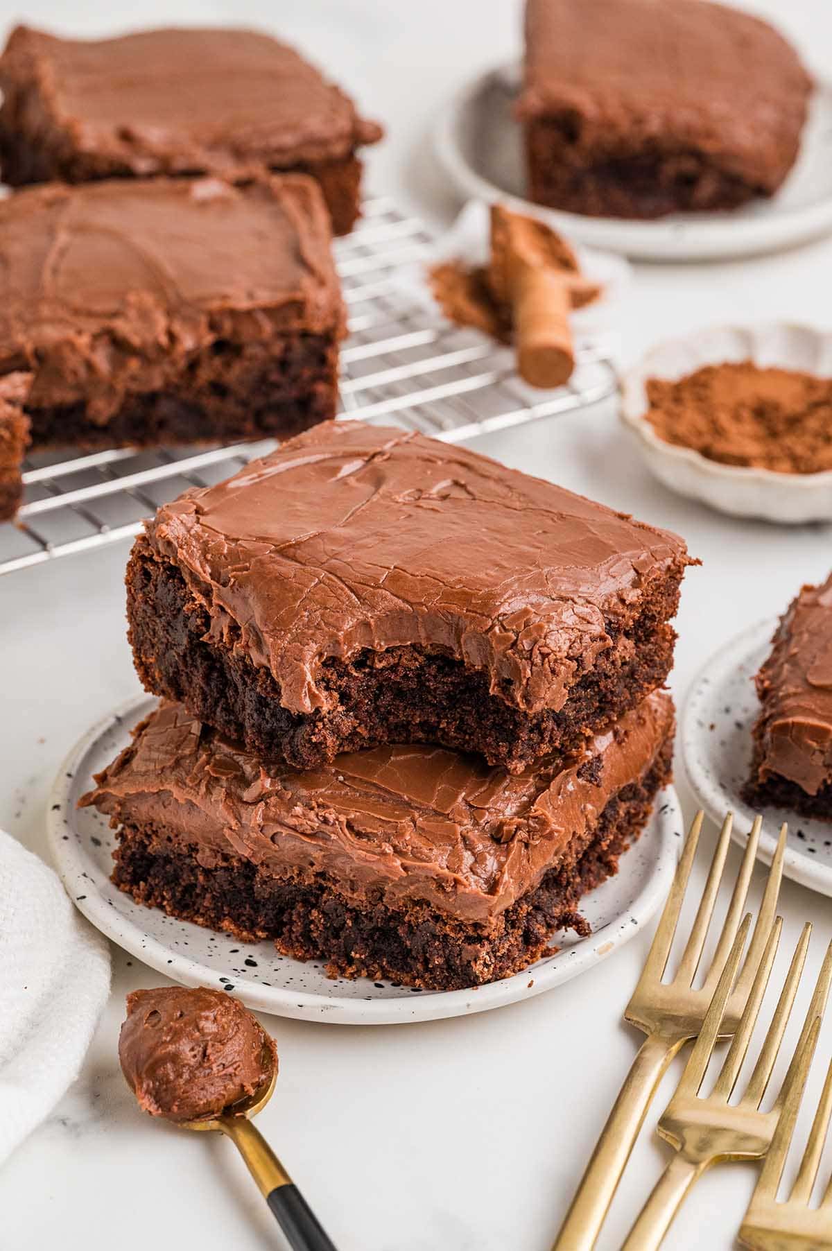 Chocolate brownies with thick frosting stacked on a plate, with more brownies in the background.