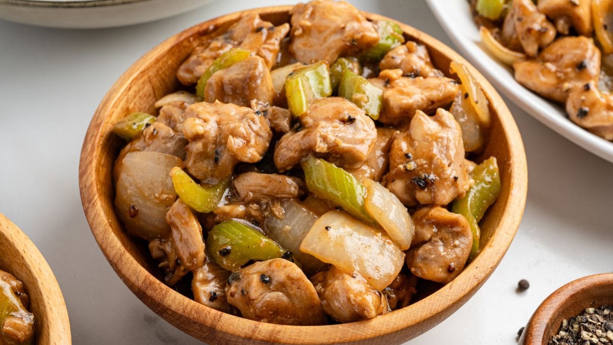 Copycat Panda Express black pepper chicken in a wooden bowl with celery and onions, with a small bowl of black pepper nearby.