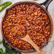 Smoked baked beans in a cast iron skillet with a wooden spoon, on a wooden board with a sliced jalape&ntilde;o and fresh parsley nearby.