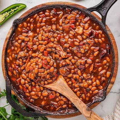 Smoked baked beans in a cast iron skillet with a wooden spoon, on a wooden board with a sliced jalape&ntilde;o and fresh parsley nearby.