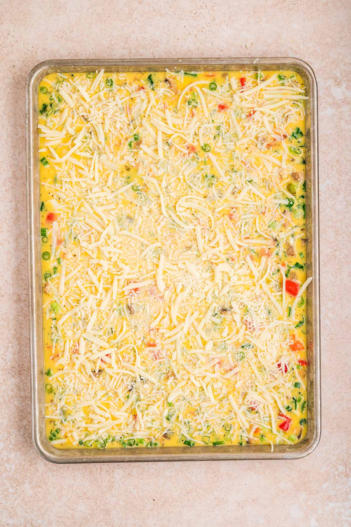 Sheet pan egg mixture with vegetables topped with shredded cheese before baking.