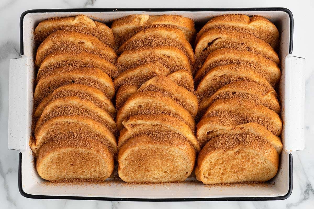 Bread slices topped with cinnamon sugar in a white baking dish ready to bake.