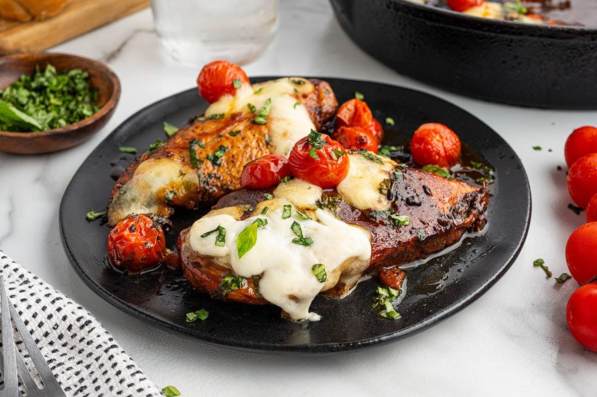 Balsamic chicken with melted of cheese served on black plate with cherry tomatoes.