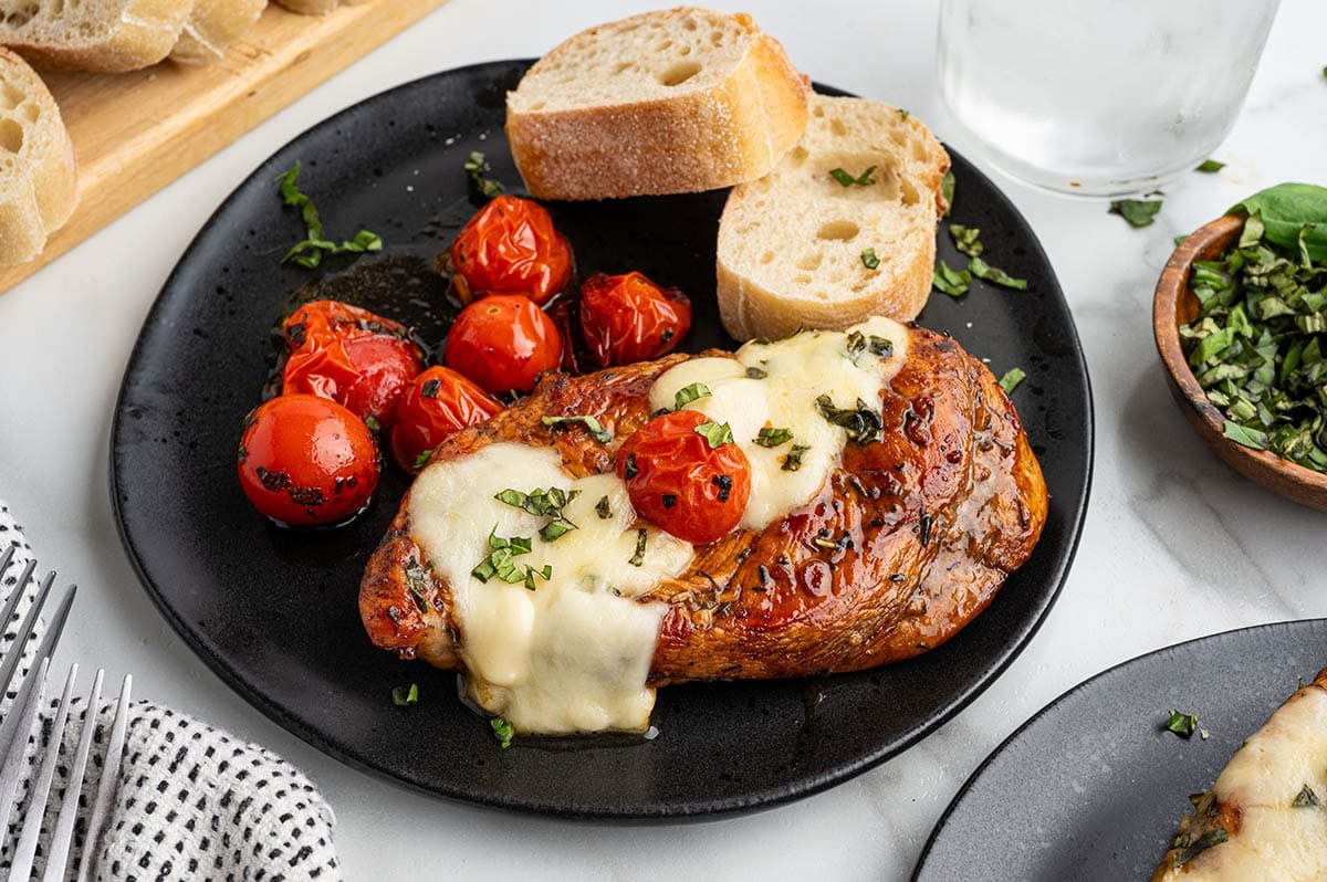 Balsamic chicken breast topped with cheese and herbs served with black plate with cherry tomatoes and slice of bread.