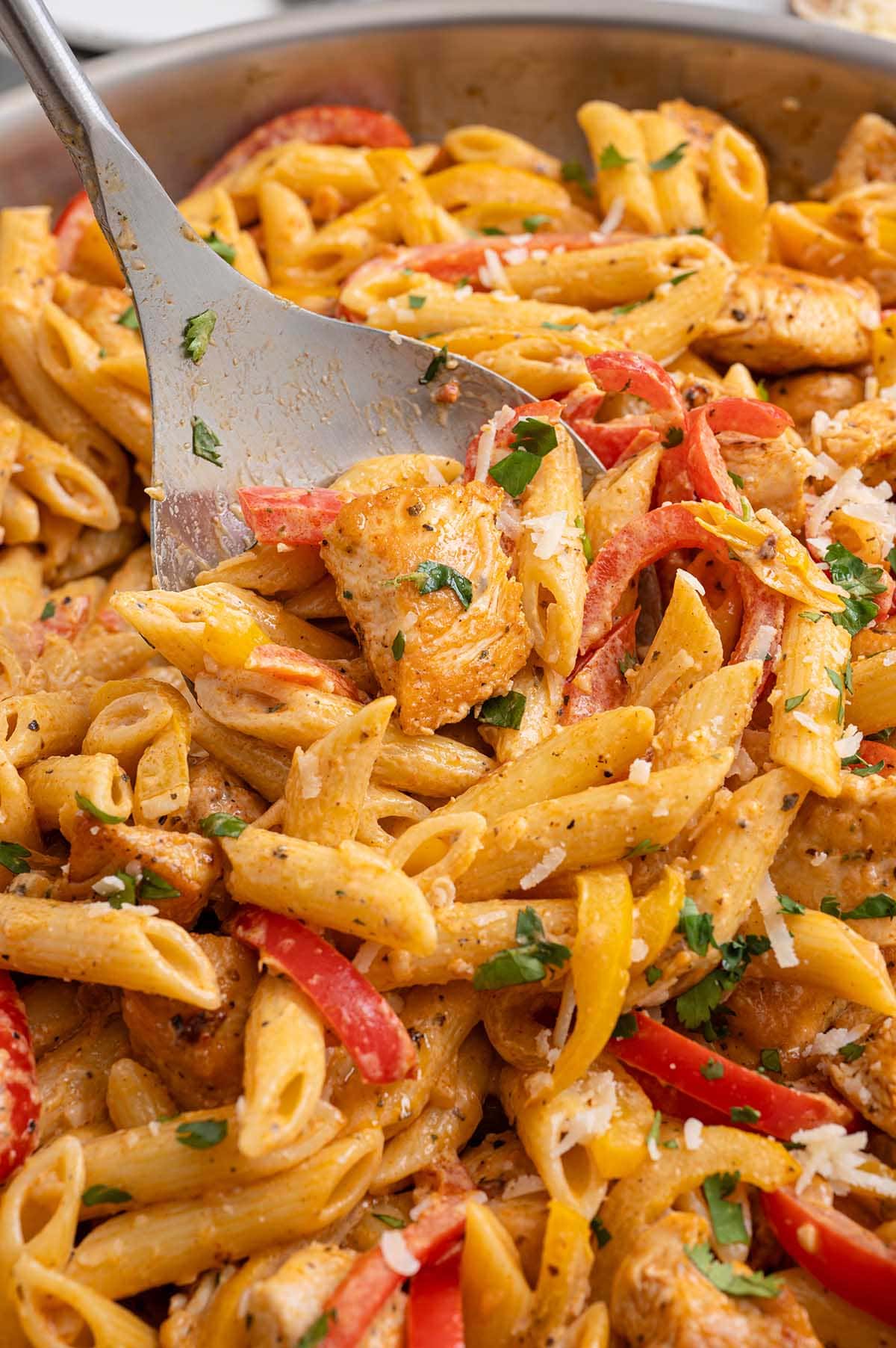 Close-up of a spoon scooping Cajun chicken pasta with bell peppers, parmesan and fresh parsley from a skillet.
