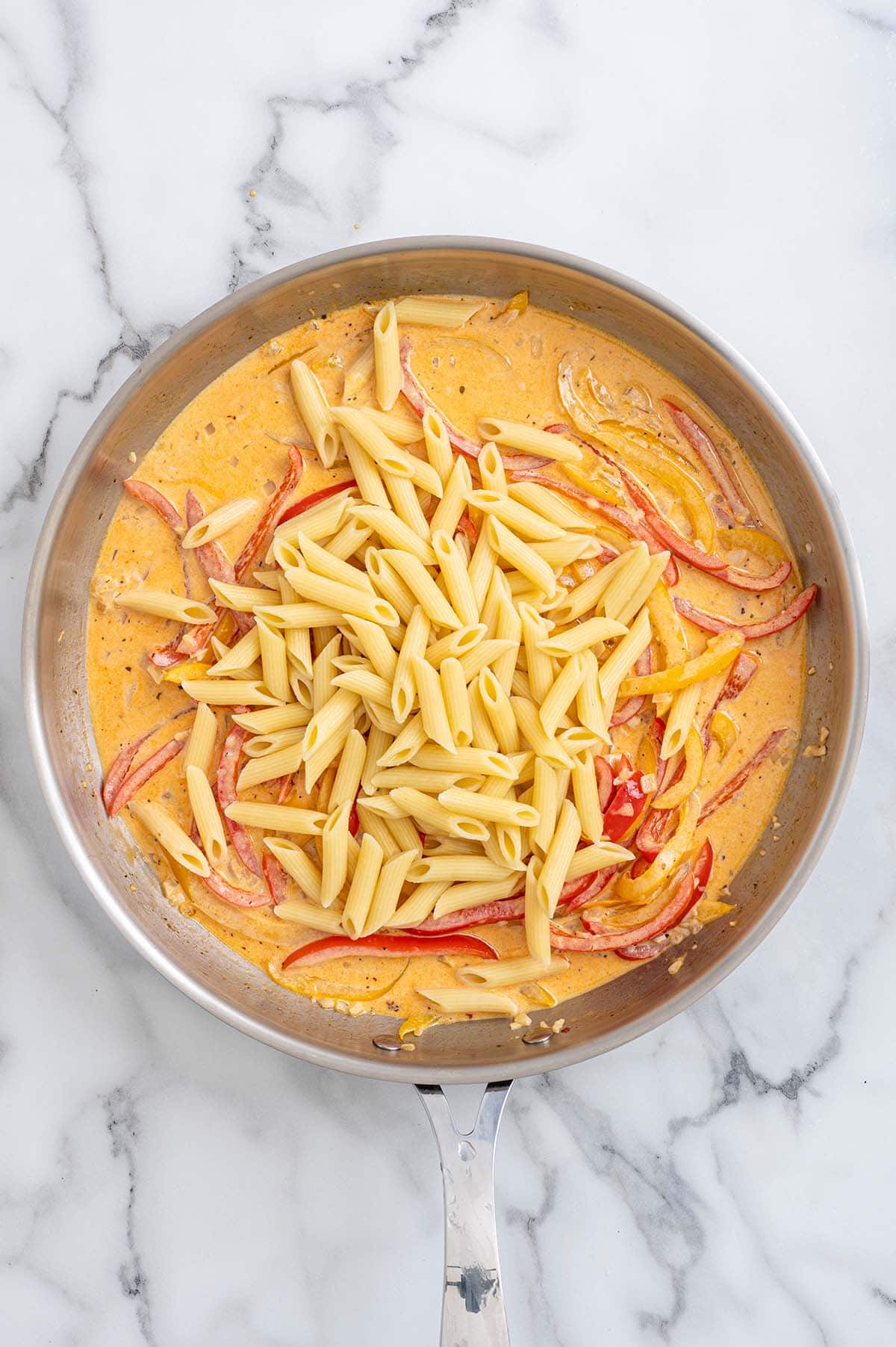 Penne pasta added to Cajun cream sauce with bell peppers in a skillet.