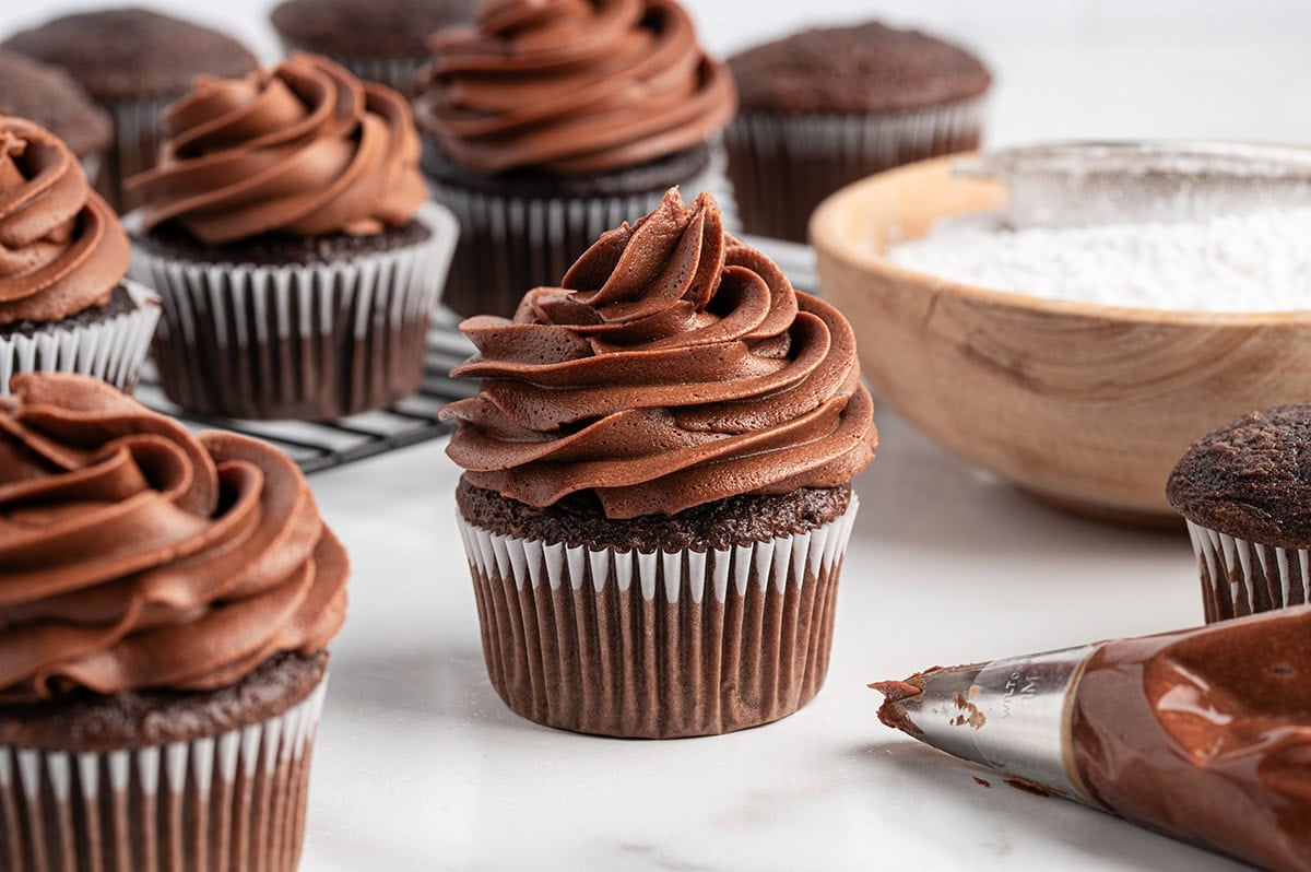 Chocolate cupcakes swirled with chocolate buttercream frosting with a piping bag and bowl of powdered sugar in the background.