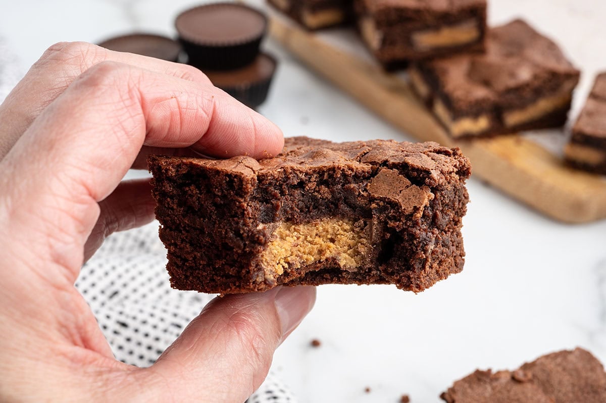 A hand holds a fudgy chocolate brownie with a bite that reveals gooey peanut butter cup filling.