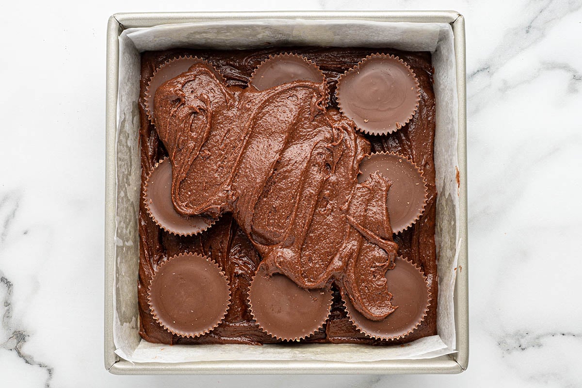 Square pan holds chocolate brownie batter with peanut butter cups pressed in and swirled chocolate on top.