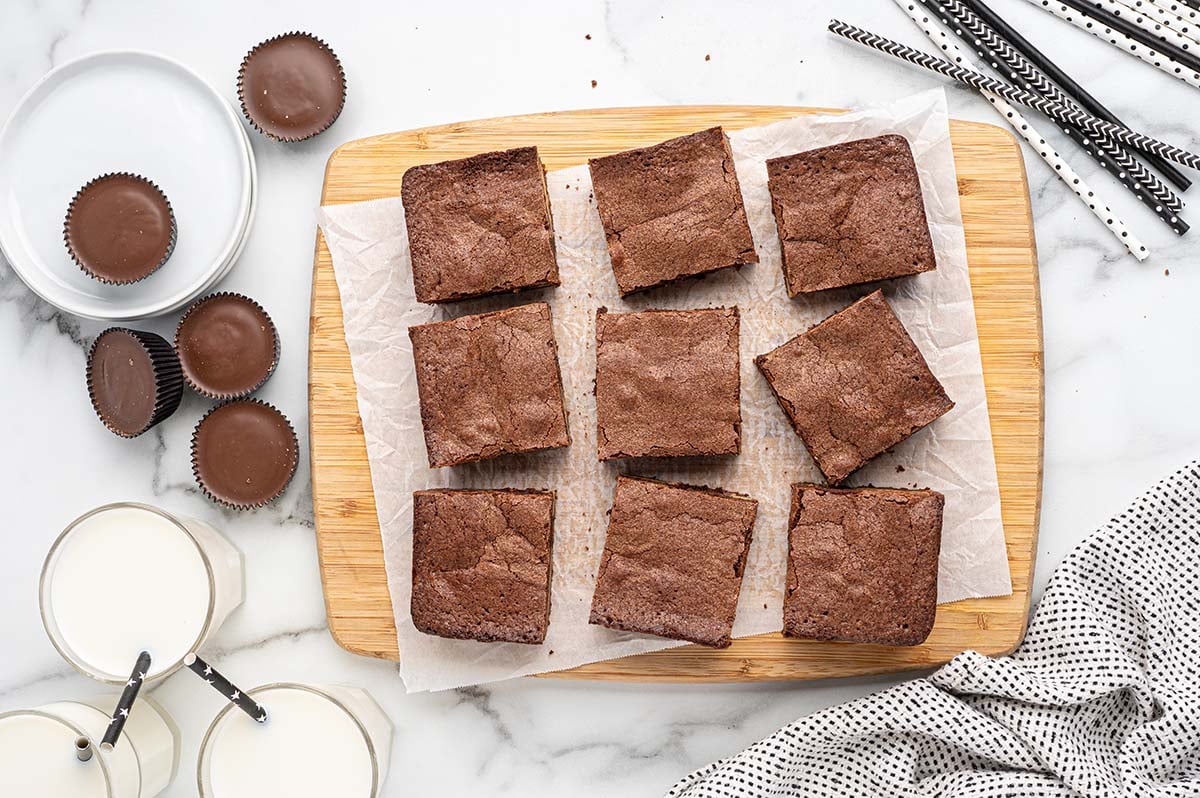 Wooden board holds brownie squares topped by peanut butter cups. White plate overflows with more. Striped straws in milk glasses complete the sweet spread.