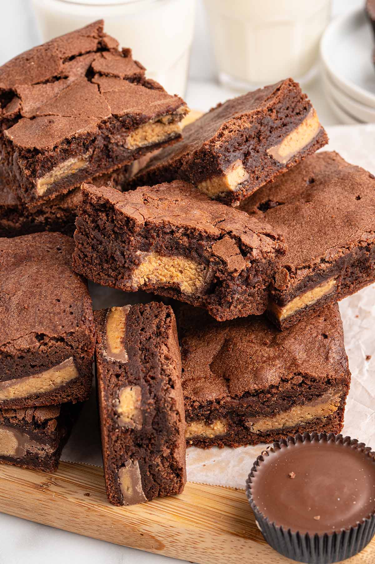 Stack of fudgy chocolate brownies filled with peanut butter cups sits on a wooden board next to milk glasses.