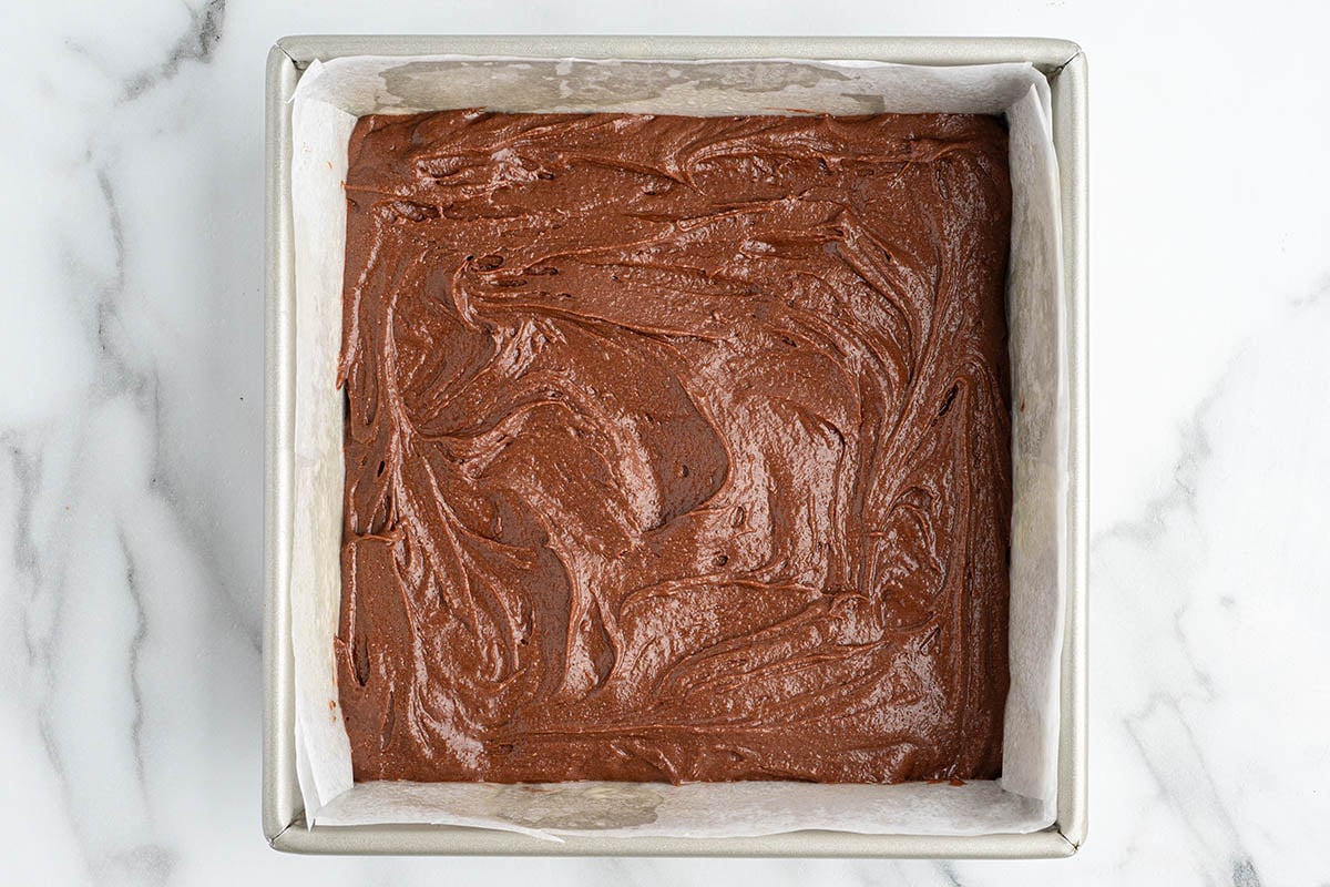 Thick chocolate brownie batter swirls in a parchment-lined square pan on white marble.