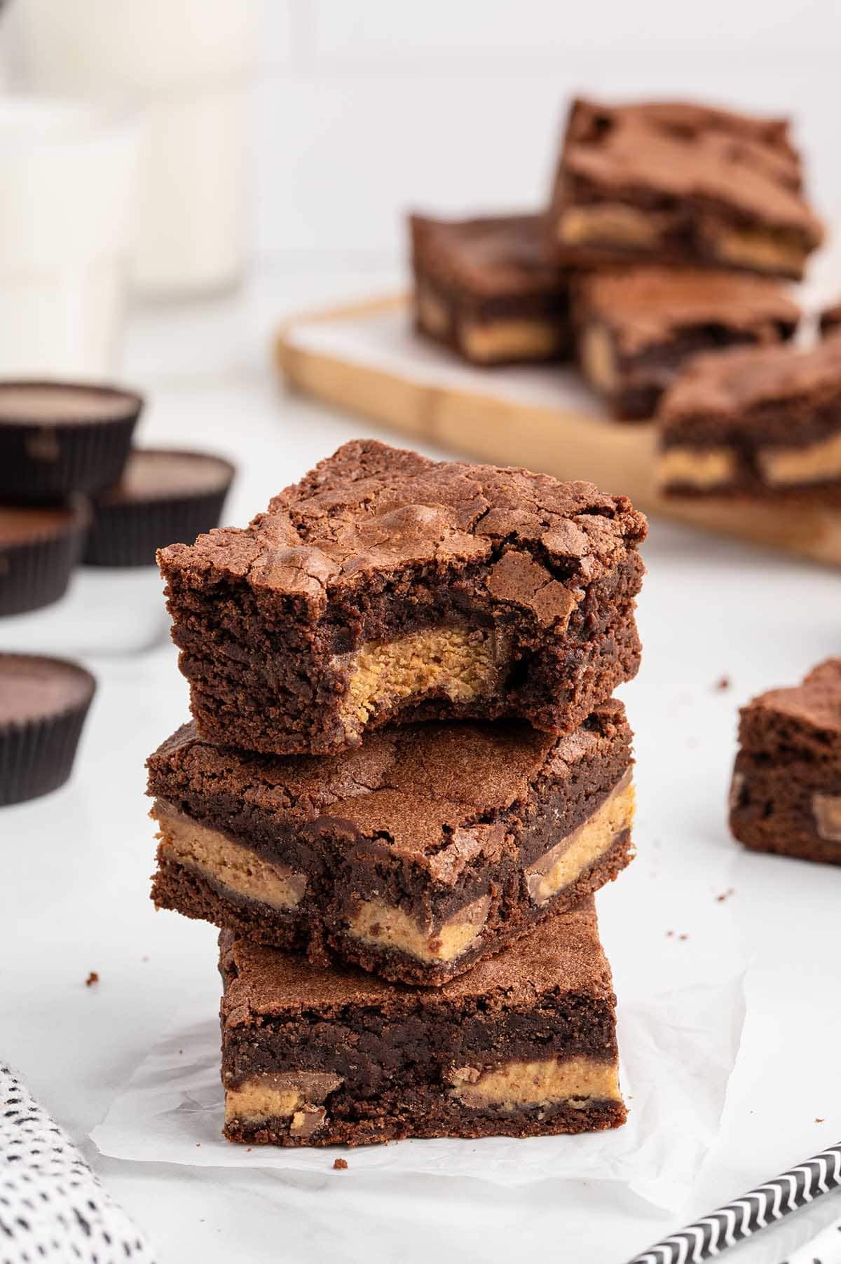 Three layered chocolate brownies with peanut butter filling exposed in a bite, next to peanut butter cups and milk.