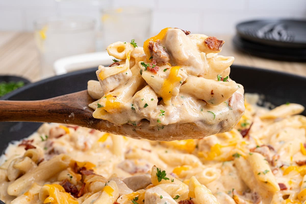 Wooden spoon scooping creamy crack chicken penne with cheddar cheese and bacon from a skillet.